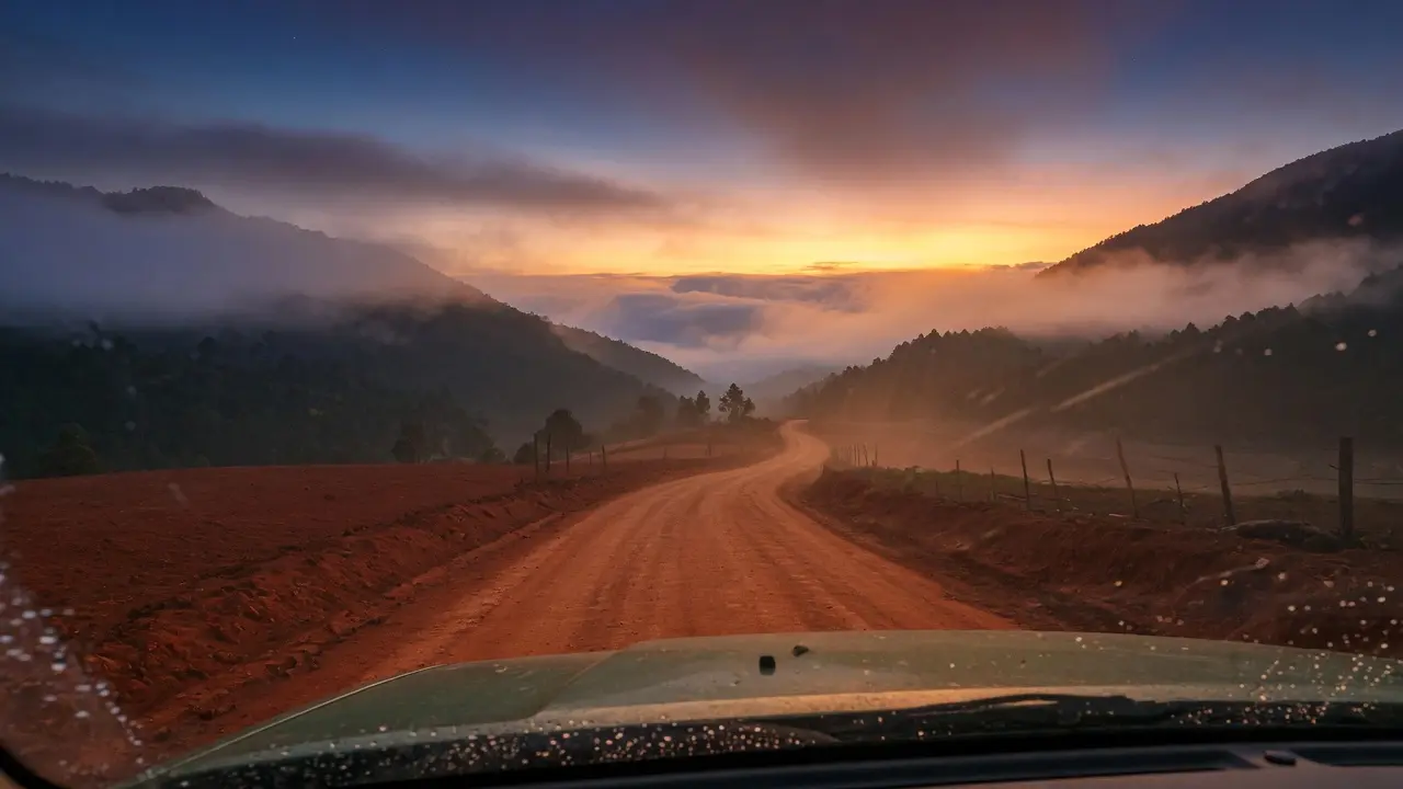 Camino rural guatemalteco al amanecer visto desde adentro de un vehículo a través del parabrisas, la carretera se pierde en la distancia entre montañas con neblina, la luz dorada del amanecer apenas asoma en el horizonte, el interior del vehículo en sombra oscura contrasta con la luz que viene, composición amplia 16:9, sin figuras humanas, atmósfera de camino largo y dolor que llega en su propio tiempo